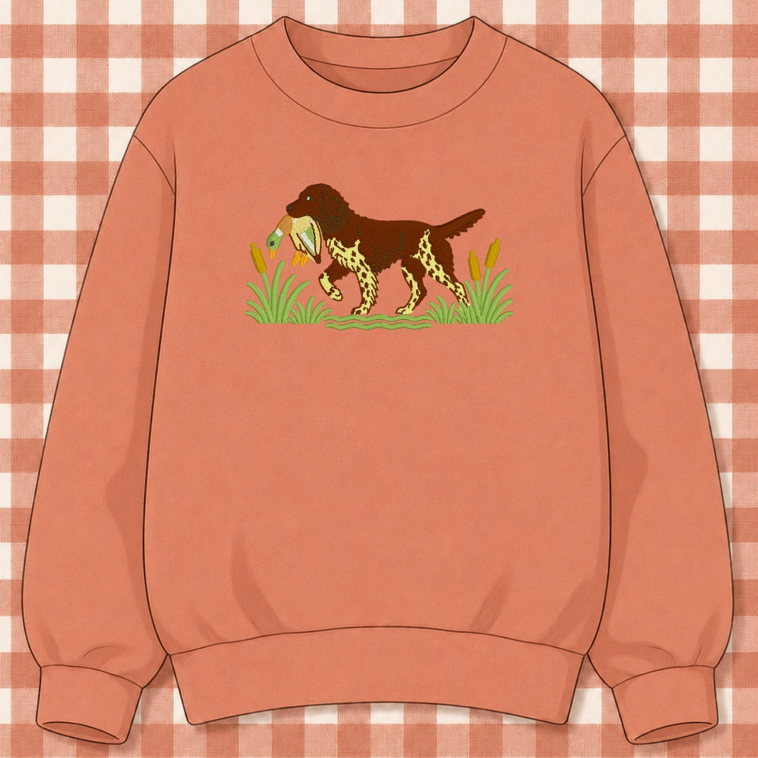 Terracotta crewneck sweatshirt with an embroidered brown and cream hunting dog carrying a mallard duck among cattails
